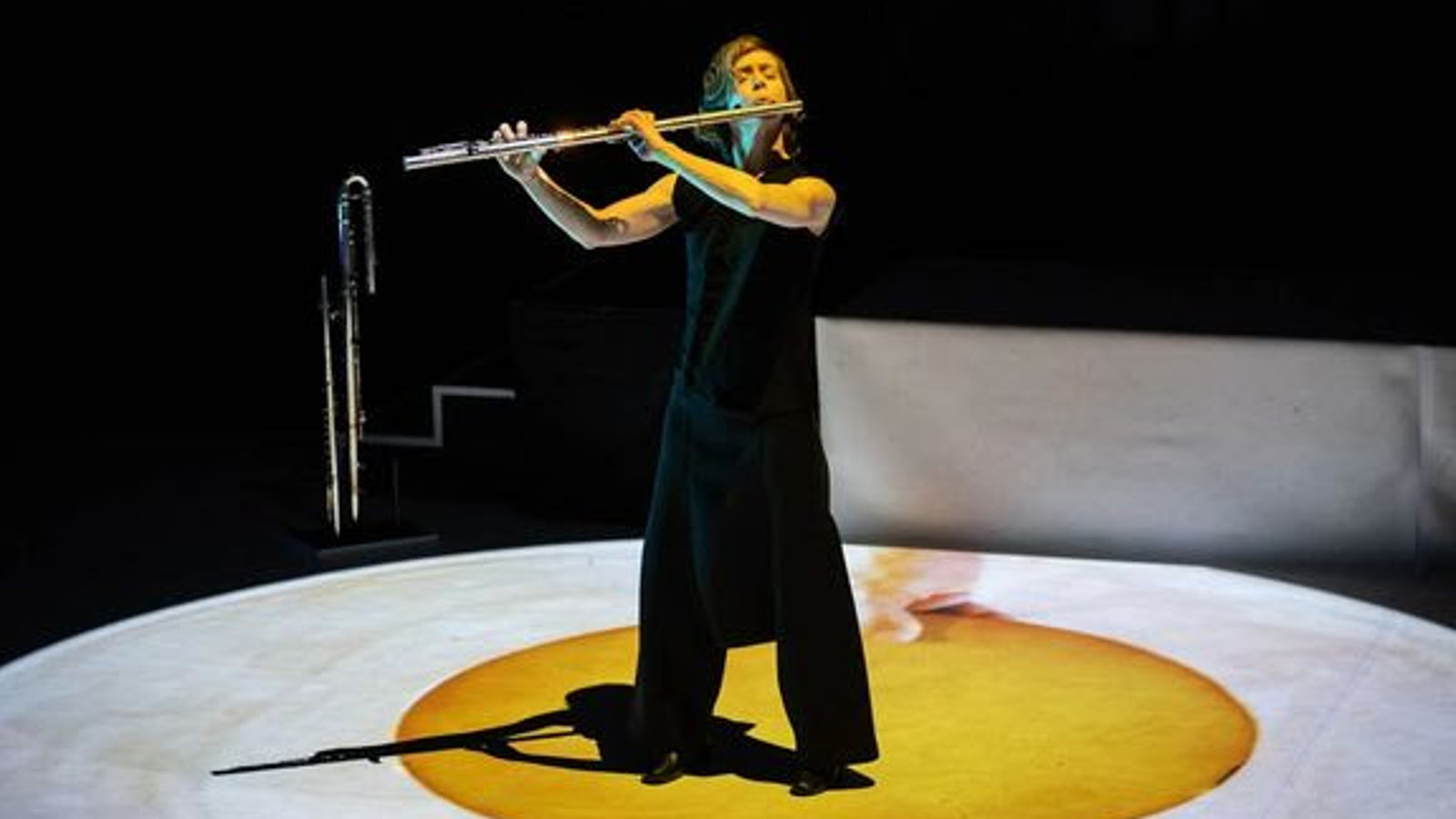 Kuvassa on Claire Chaise soittamassa huilua seisten ja hänellä on musta hihaton musta mekko sekä pitkät housut. Hän seisoo keltaisen, pyöreän maton päällä ja sen ympärillä on valkoinen ympärillä. Taustalla on mustaa.